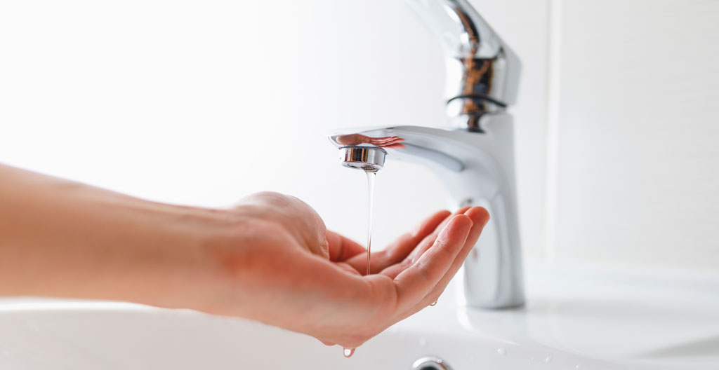 Main de propriétaire tenant un verre sous un robinet de cuisine moderne au débit d'eau anormalement faible, dans une cuisine résidentielle québécoise contemporaine bien éclairée